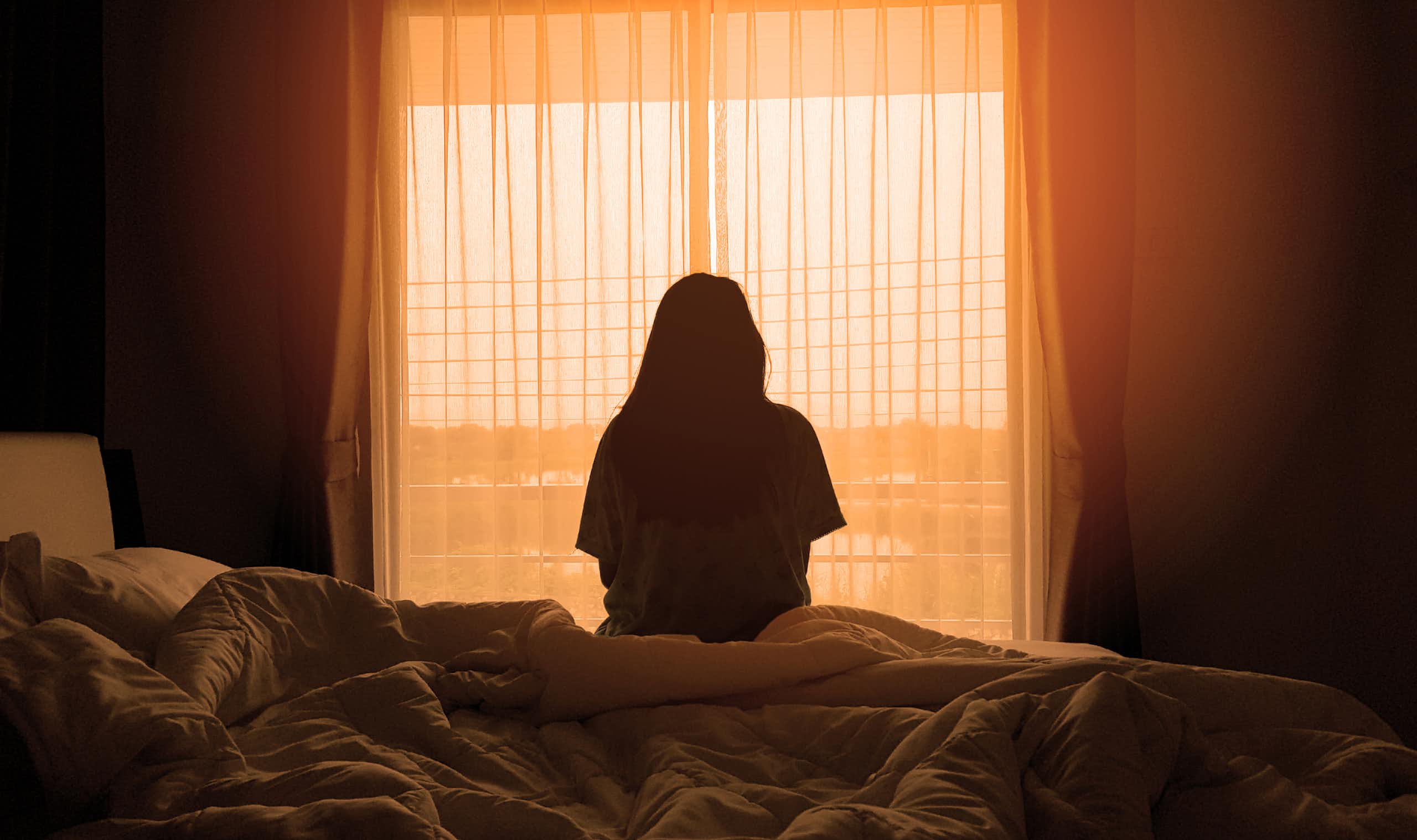 A woman sitting on the edge of a bed in a darkened room looking out of the window.