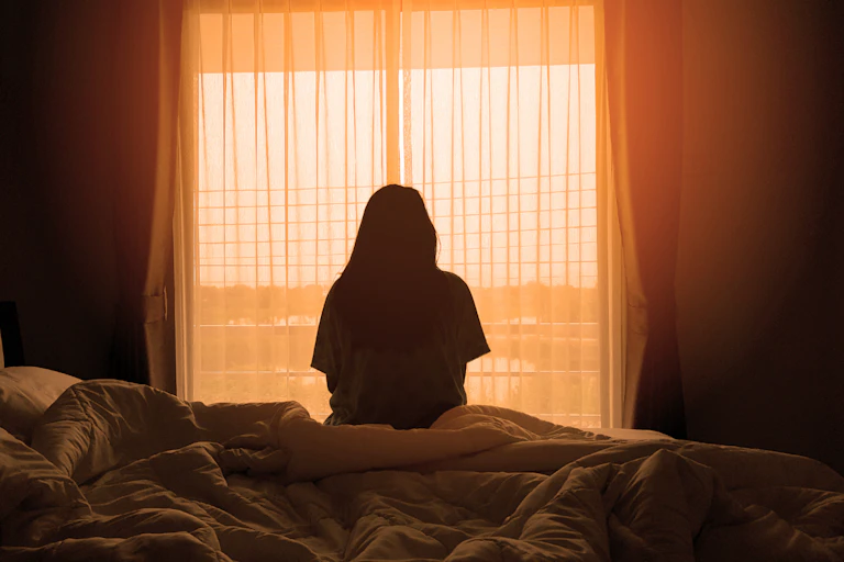 A woman sitting on the edge of a bed in a darkened room looking out of the window.