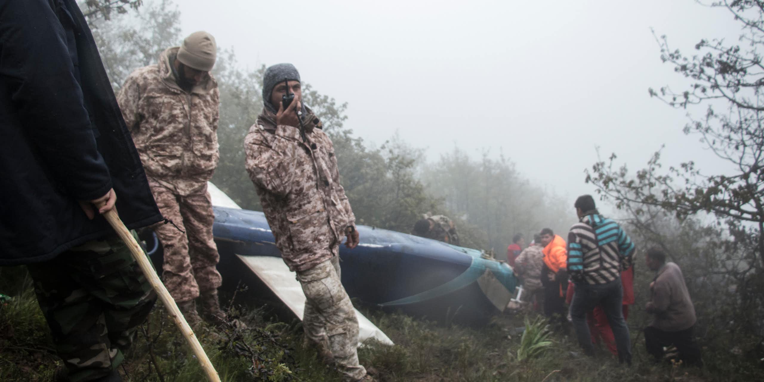A rescue worker talking into a radio in front of a helicopter wreckage. 