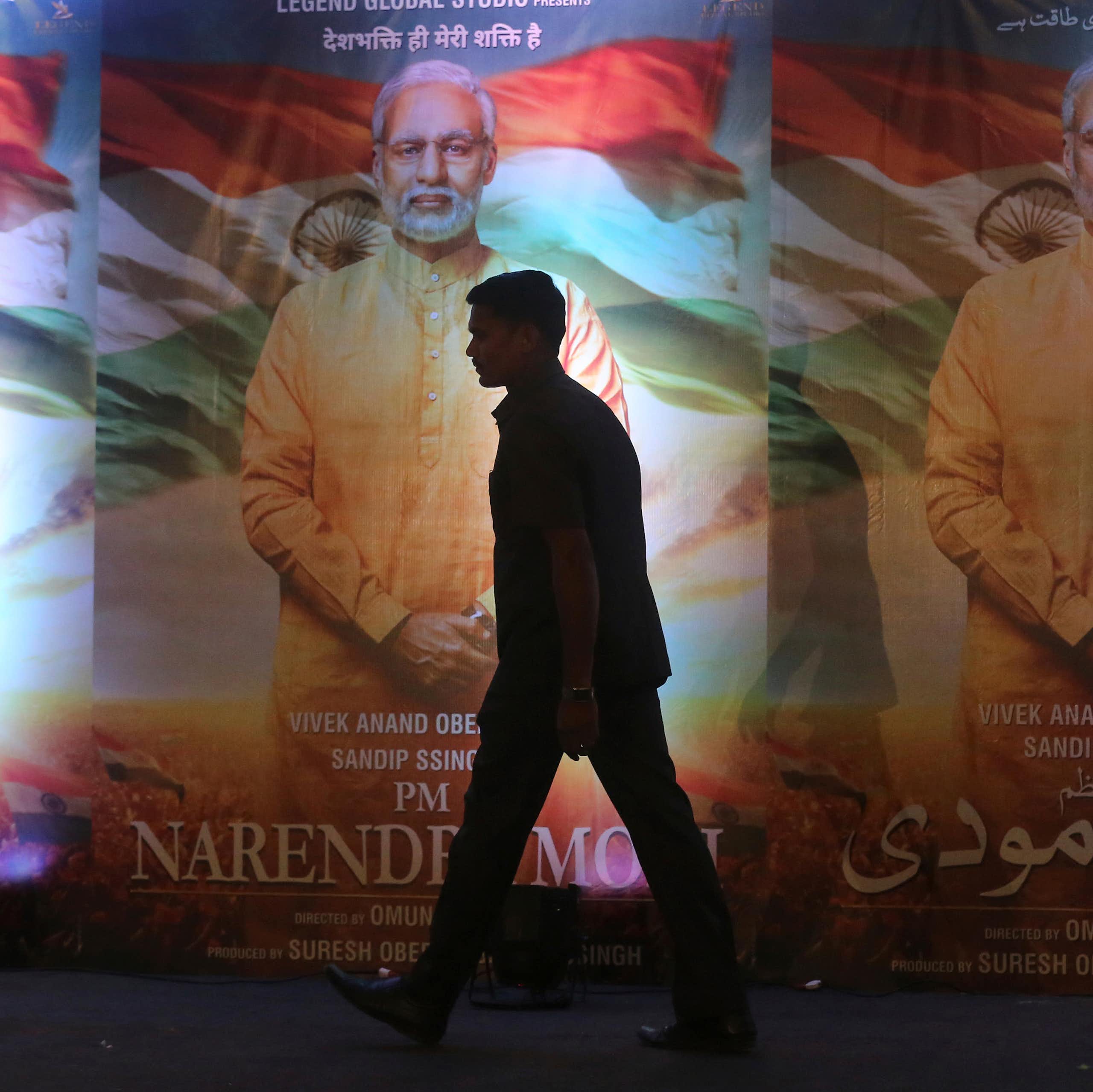 Silhouette d'un homme passant devant trois affiches identiques représentant un homme aux cheveux gris, vêtu d'une longue chemise couleur safran, avec le drapeau de l'Inde en arrière-plan.