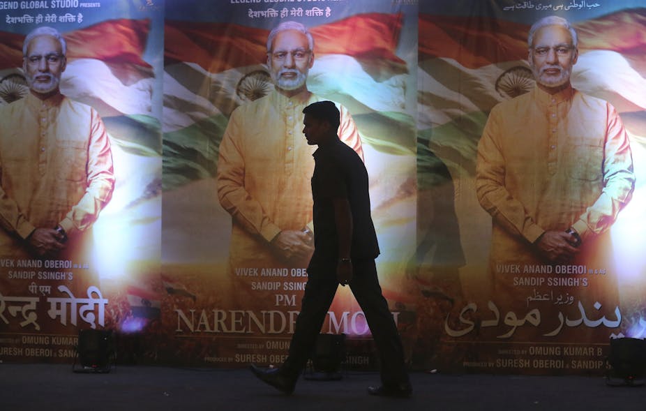 Silhouette d'un homme passant devant trois affiches identiques représentant un homme aux cheveux gris, vêtu d'une longue chemise couleur safran, avec le drapeau de l'Inde en arrière-plan.