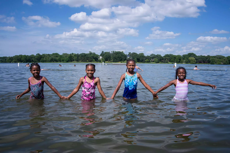 Four young girls look at the camera, waist deep in a lake, smiling and holding hands.