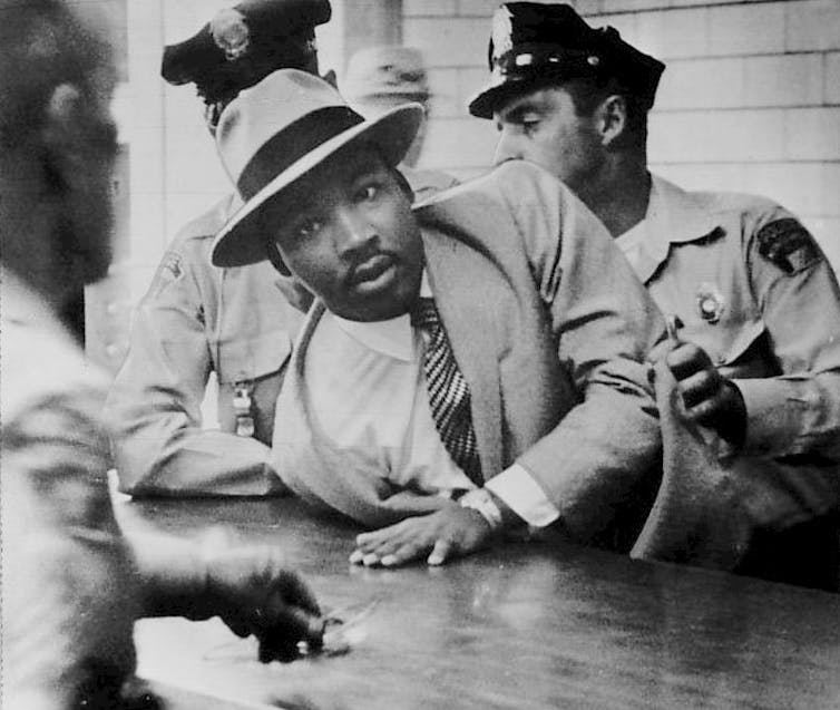 A Black man in a coat, tie and fedora is arrested by police.