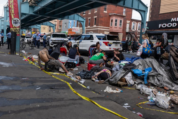 People sit on street surrounded by trash and personal belongings while police officers and vehicles are in background