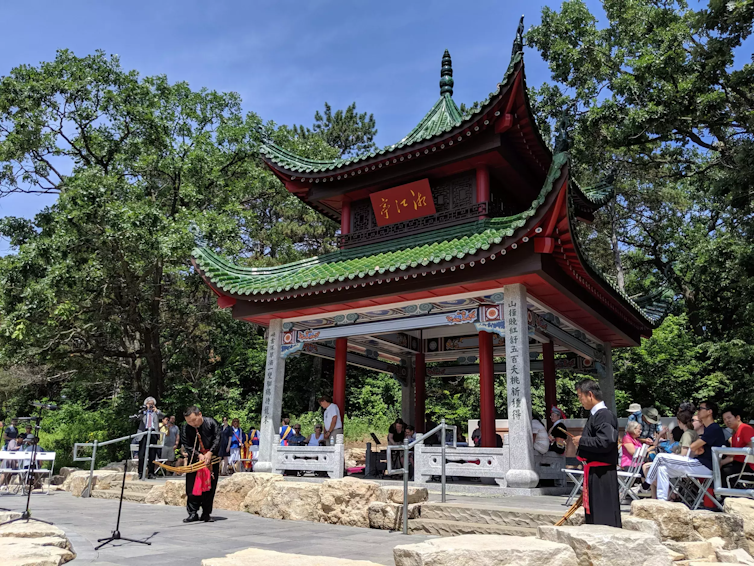 People conduct a ceremony at a large open Chinese pavilion with an upturned roof. Several people play musical instruments.