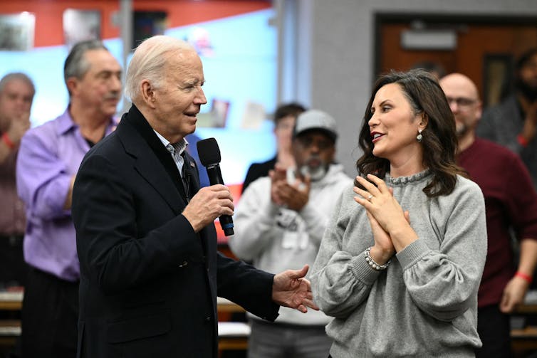 A white-haired man in a blue jacket, holding a microphone, talks to a woman with dark hair in a gray blouse in front of a few people.