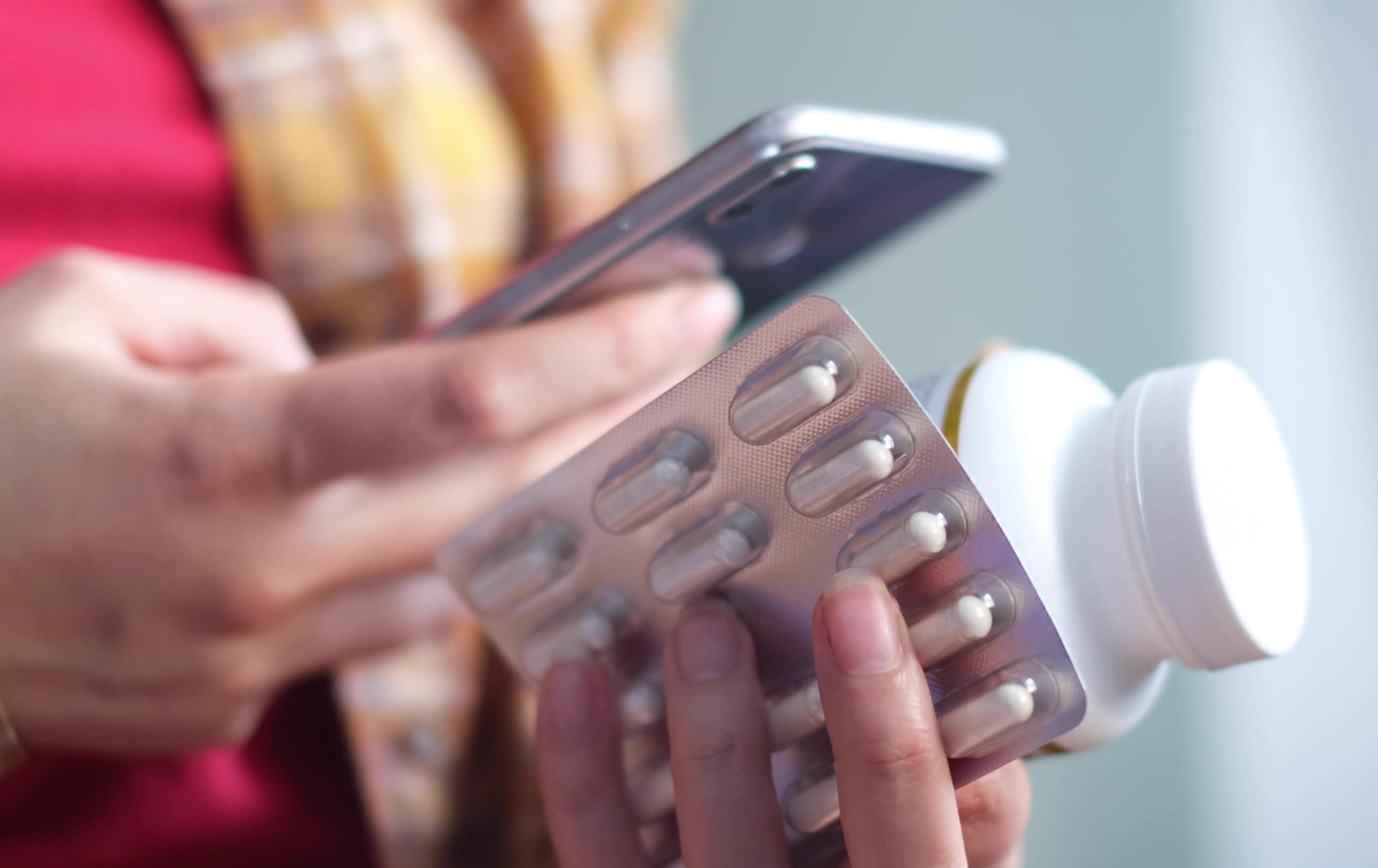 Close-up of a woman's hands holding a smartphone and some medications.