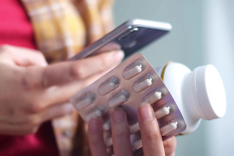 Close-up of a woman's hands holding a smartphone and some medications.
