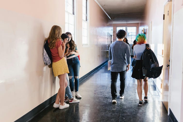 High school students walk in a school hallway. A group also leans against a wall talking.