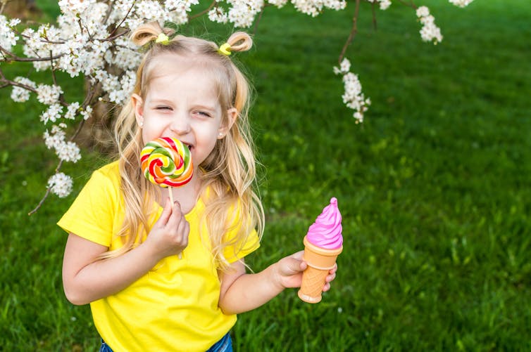 girl in yellow top licks large lollipop while holding a pink icecream