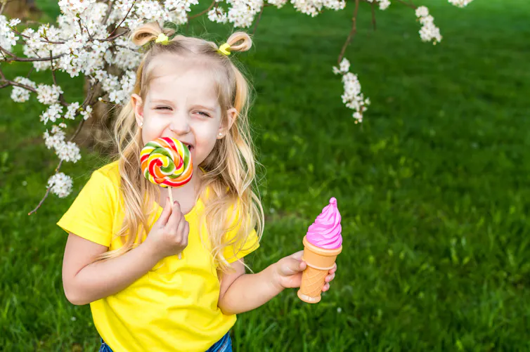 girl in yellow top licks large lollipop while holding a pink icecream