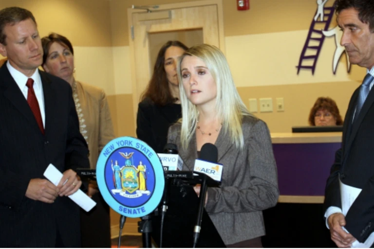 Two men in suits flank a blond-haired woman standing at a lectern.