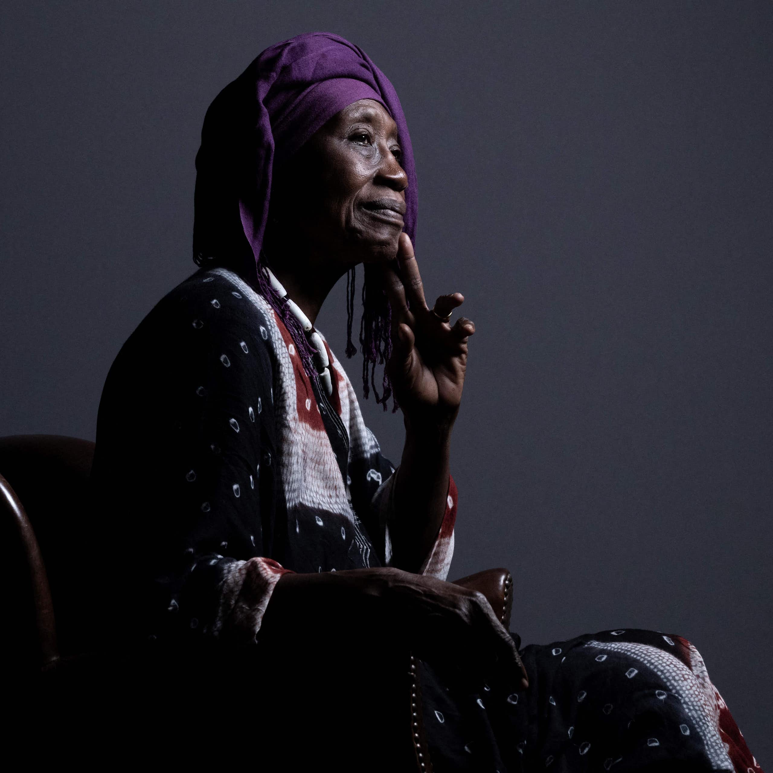 An elderly African woman sits in a chair posing for a portrait in profile, dressed in traditional African attire with a hand to her chin and smiling slightly.
