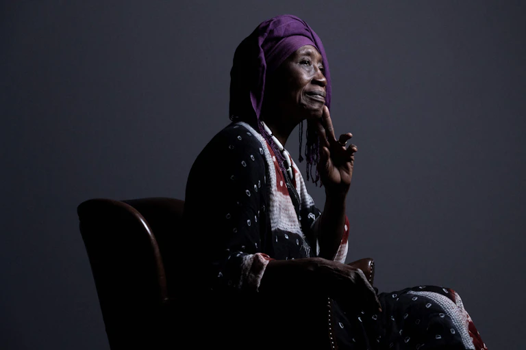 An elderly African woman sits in a chair posing for a portrait in profile, dressed in traditional African attire with a hand to her chin and smiling slightly.
