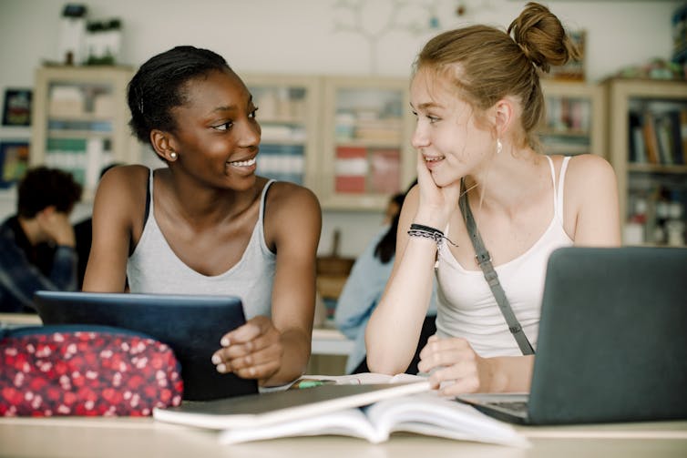 Two teen girls in white tank tops smile and chat while they're seated at a table in a classroom.