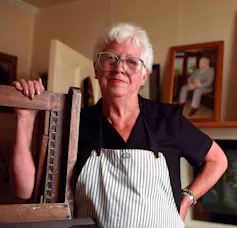 A middle-aged woman stand in her art studio with her work in the background.