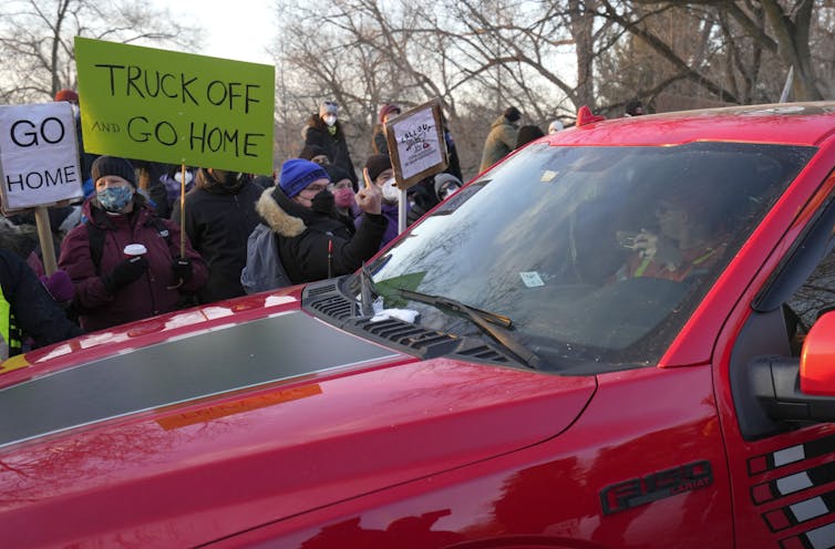 A red pickup truck amid a crowd of people waving signs. One reads Go Home.