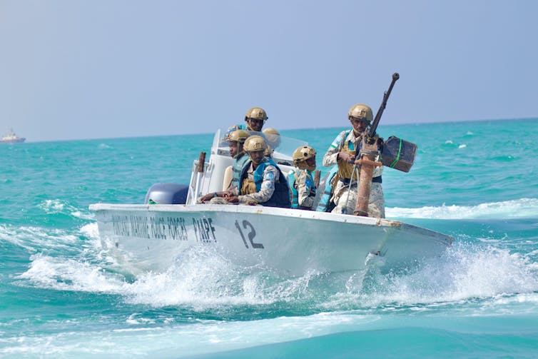 Six policemen with guns and helmets in a small boat driving through a clear blue sea.