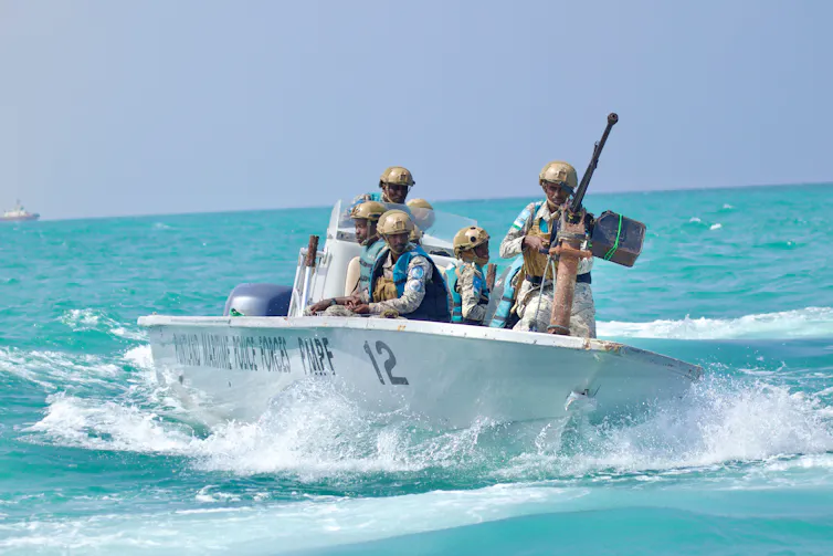 Six policemen with guns and helmets in a small boat driving through a clear blue sea.