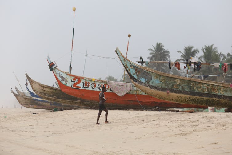 A man walking up a beach in front of a line of wooden fishing boats on a foggy day.