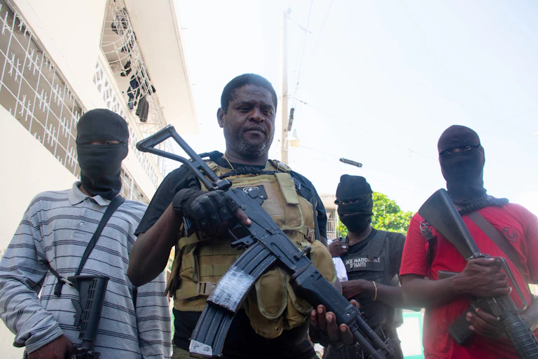 A man holding a large automatic rifle is seen flanked by men in face masks.