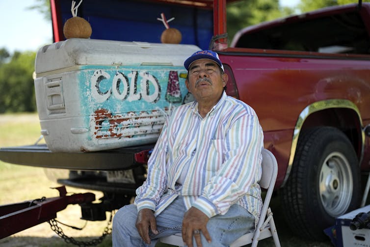 A man sits in chair closing his eyes next to a cooler on the back of a truck.