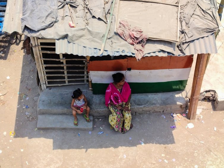 A woman and young girl sit in shade outside a small building with slats in the doors and a precarious looking roof.