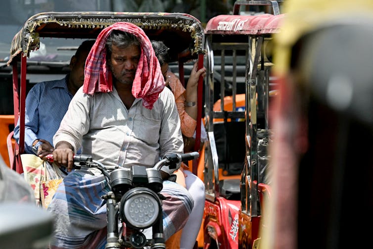 A older man driving an electric rickshaw with two passenger sits in direct sunlight in traffic on a hot day. He had a bandana over his head for shade.