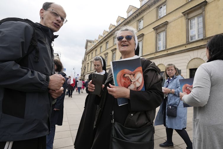 A woman wearing sunglasses and a nun's habit holds a picture of a child in the womb while talking to a man on the street.