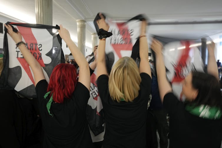 Three women in black t-shirts, seen from the back, hold up scarves with a red, white and black design.