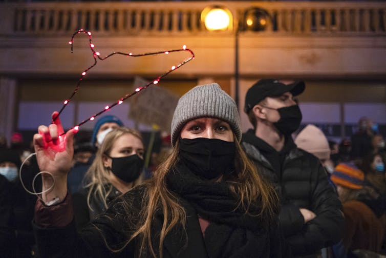 Women and men in face masks and coats stand in an area illuminated by a streetlight.