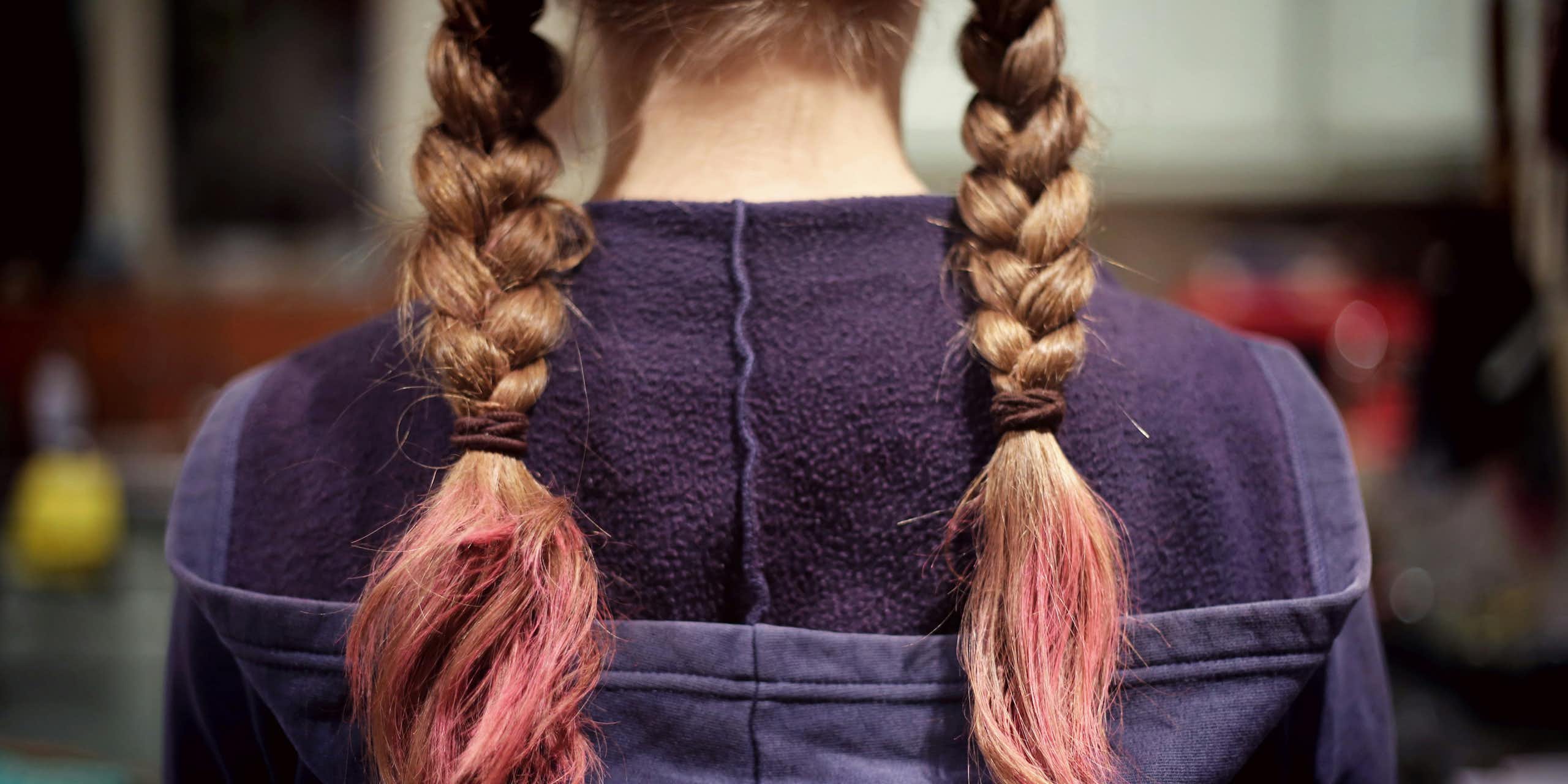 A young person stands with their back to the camera. They have a hoodie jumper on and blonde braided pigtails died pink at the ends. 
