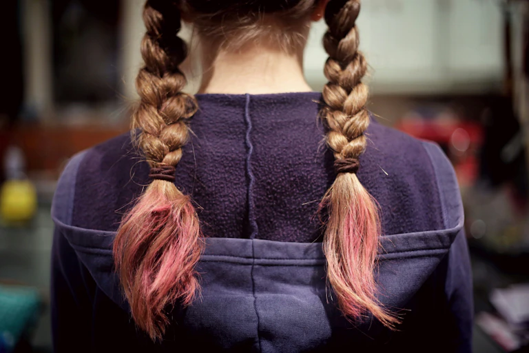 A young person stands with their back to the camera. They have a hoodie jumper on and blonde braided pigtails died pink at the ends.