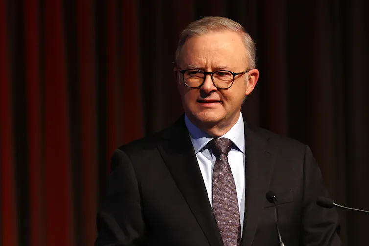 A man in a suit speaks at a lectern in front of a dark red curtain