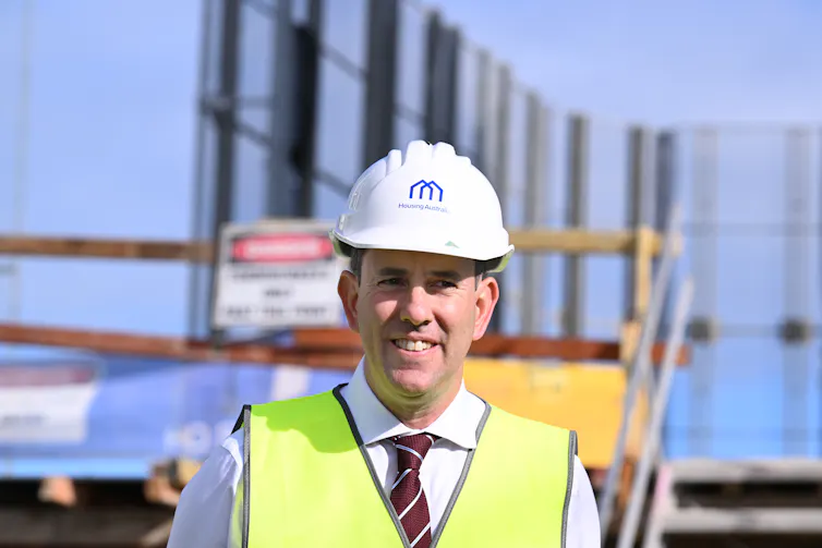 A man in a shirt, tie and high-vis vest wears a hardhat and smiles