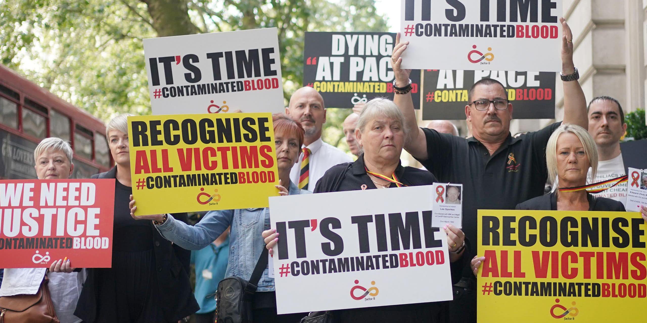 A group of people holding protest signs in red, yellow, black and white colours that say 'it's time' and 'recognise all victims' with the hashtag Contaminated Blood