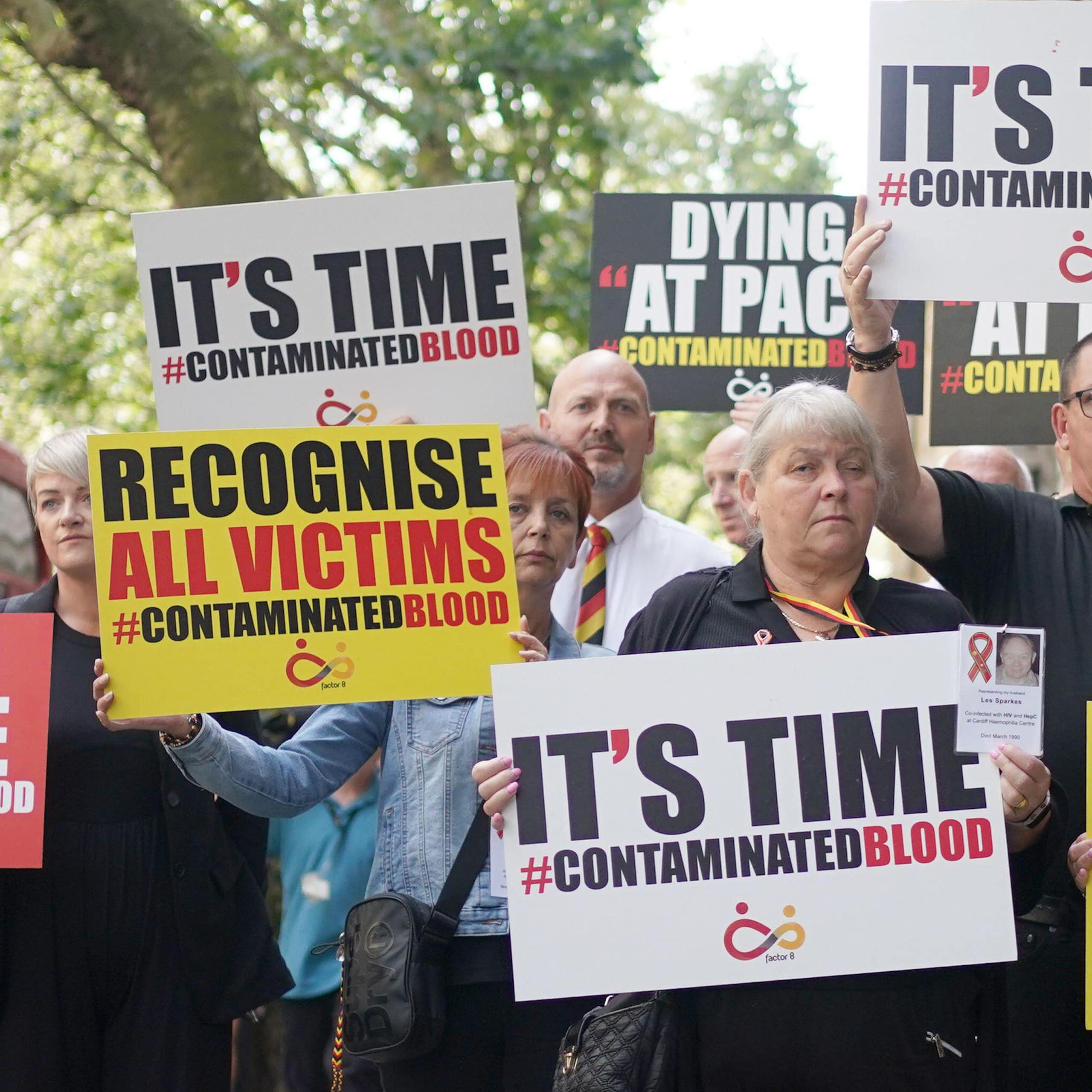A group of people holding protest signs in red, yellow, black and white colours that say 'it's time' and 'recognise all victims' with the hashtag Contaminated Blood