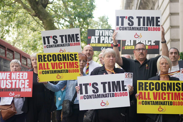 A group of people holding protest signs in red, yellow, black and white colours that say 'it's time' and 'recognise all victims' with the hashtag Contaminated Blood