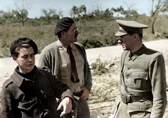Two men in civilian clothing speak to a solider in a field.