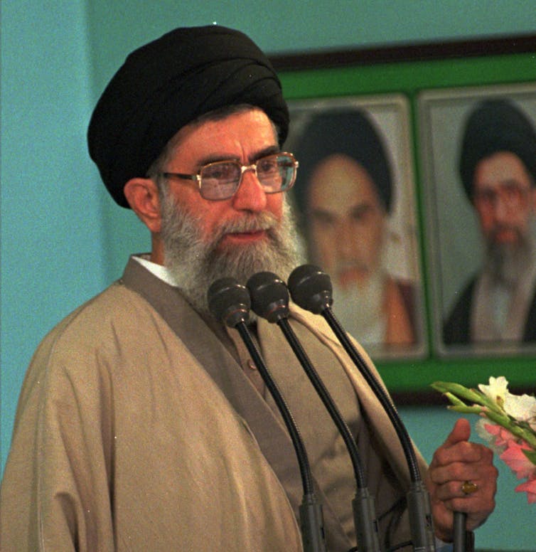 A bearded man wearing a black turban and a long robe speaks on microphones while holding white flowers in one hand. On the wall next to him are photographs of two men, also wearing black turbans.