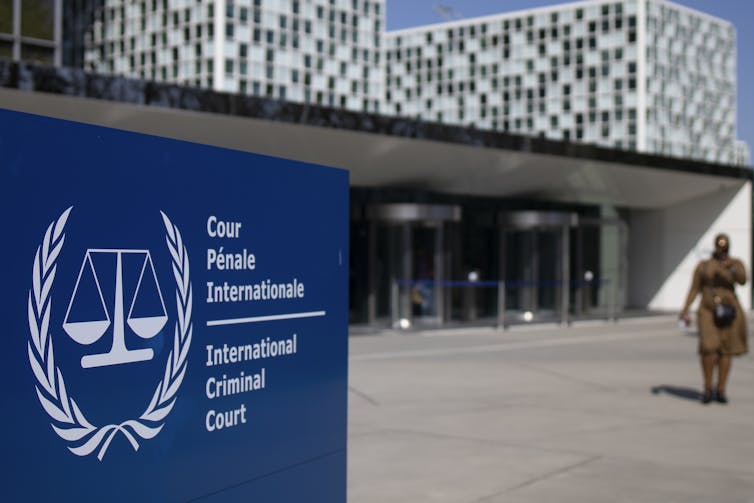 A blue sign reads International Criminal Court in front of a modern building. Someone is walking towards the building to the right of the sign.