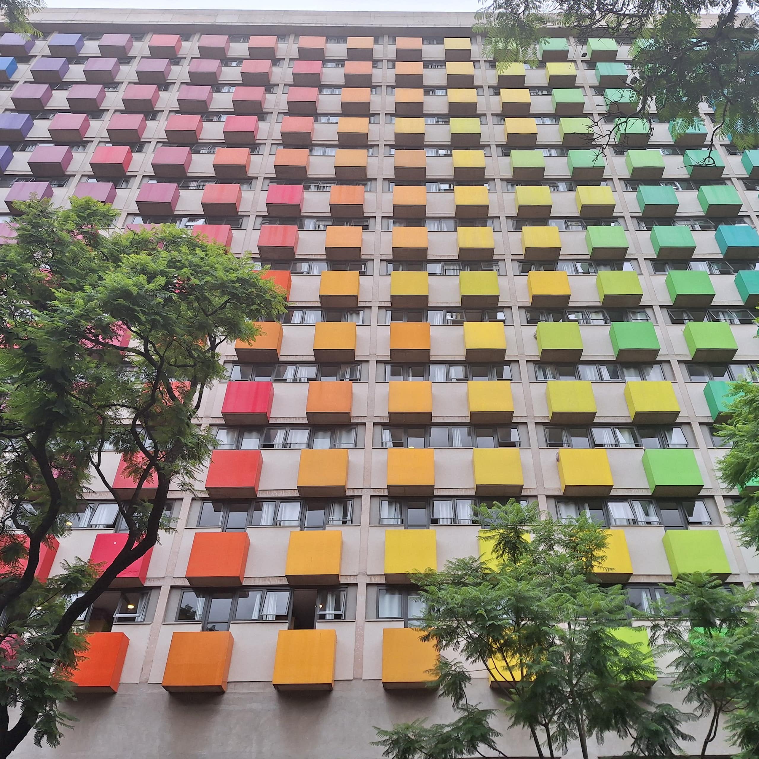 A high-rise building with blocks of colour on the exterior of each unit, forming a rainbow of colours, trees in the foreground. 
