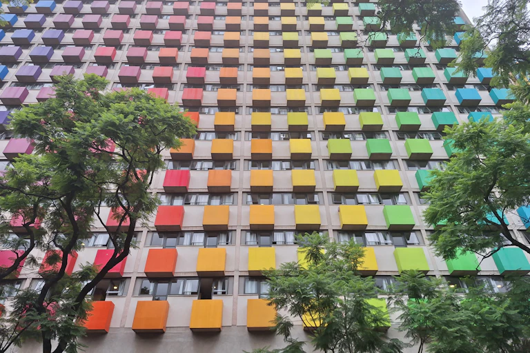 A high-rise building with blocks of colour on the exterior of each unit, forming a rainbow of colours, trees in the foreground.