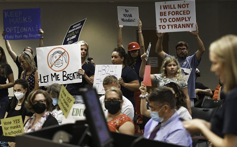 People with signs protesting at a meeting; one sign reads 'I will not be forced to comply to tyranny.'