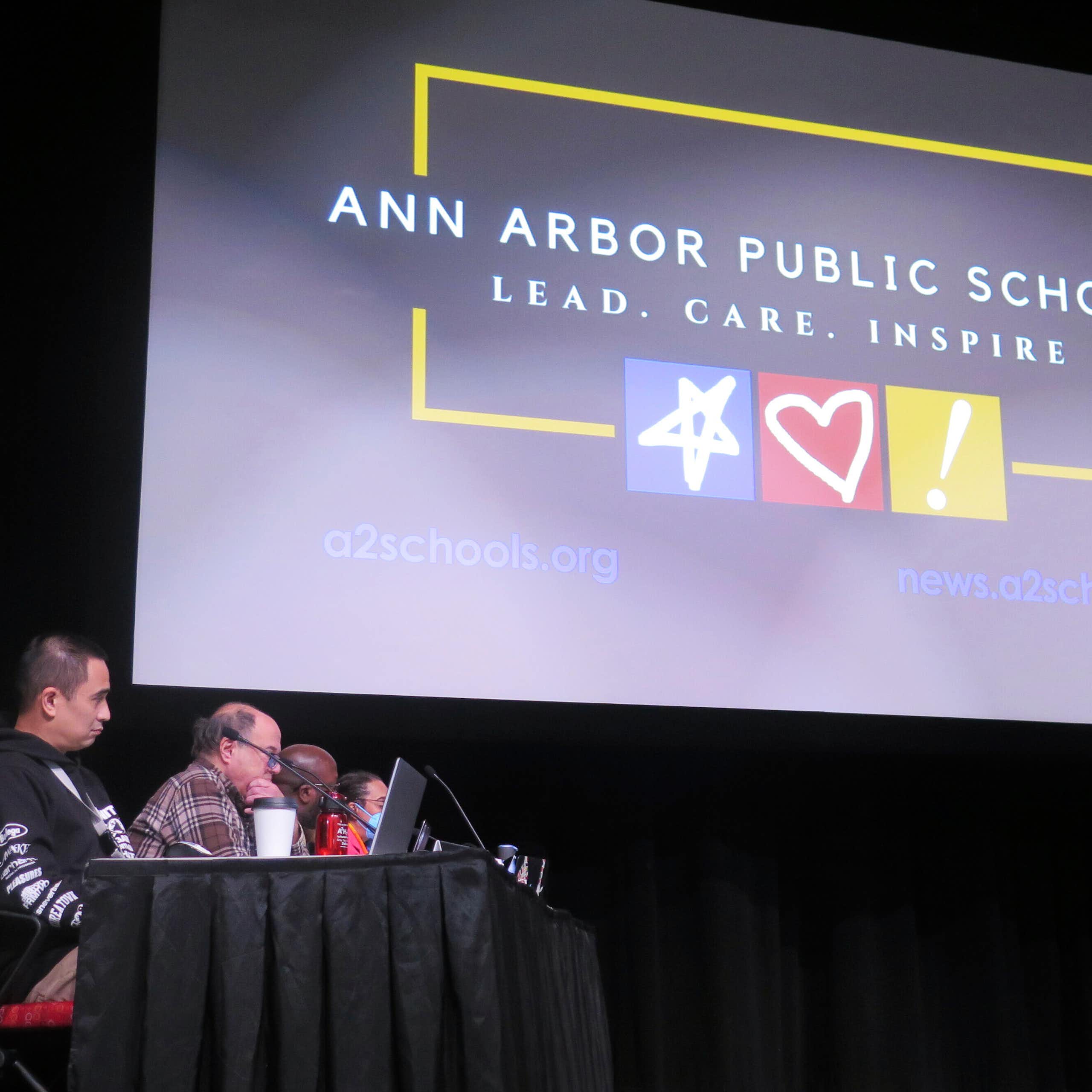 At least four people sitting at a skirted table, with the sign 'Ann Arbor Public Schools' above them.