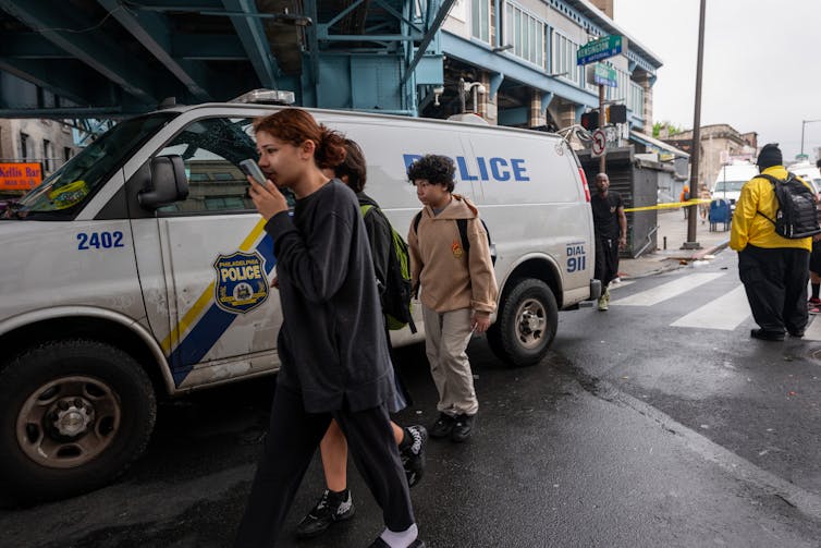 People walk past a police van in street underneath elevated train