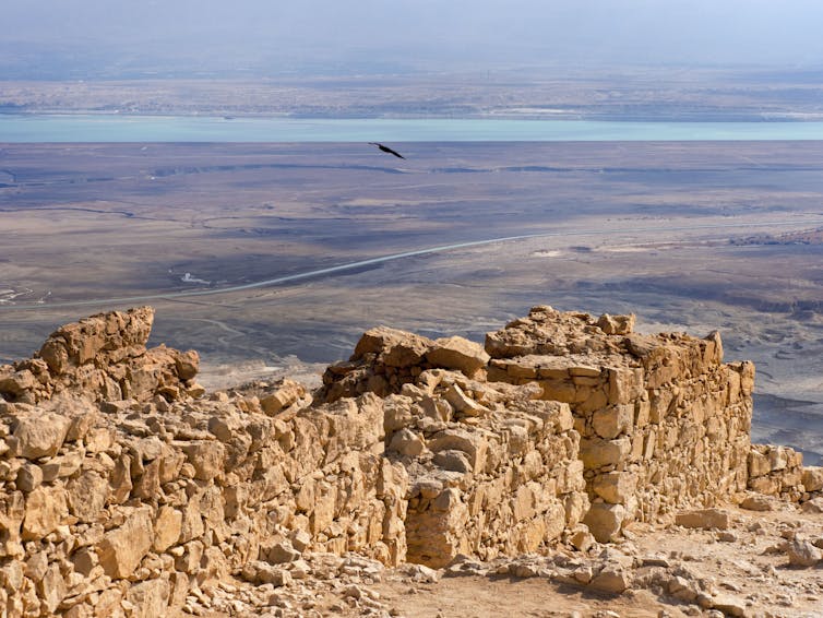 Large brown ruins overlook a large desert and a small blue body of water in the distance.