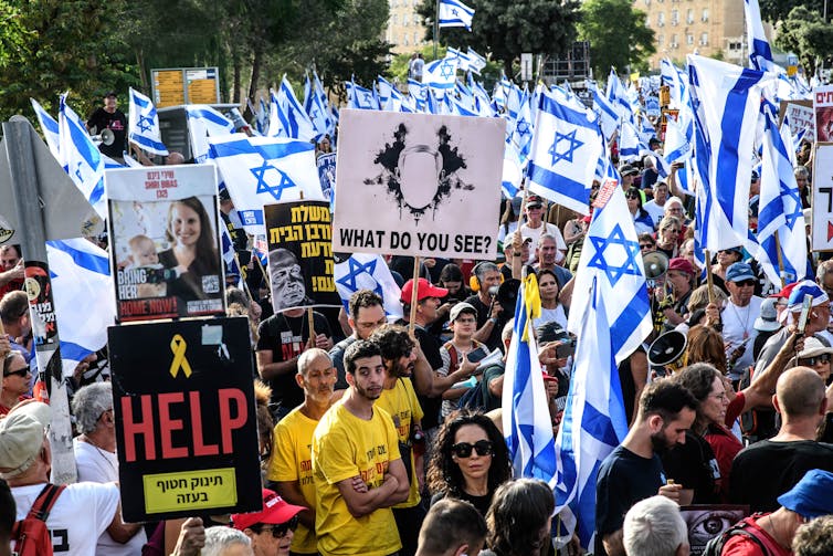 Una gran multitud de personas sostiene banderas israelíes y carteles en hebreo que también tienen la palabra 