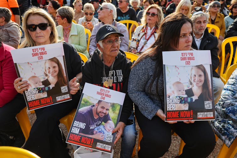 Three women sit on yellow chair and hold posters with photos of a young woman holding a baby and a man posing with a baby. The posters say 'Bring her home now!' and 'Bring him home now!'