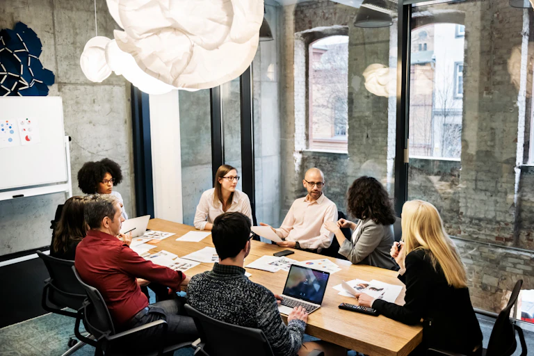 A group of young people, male and female, of different ethnicities are sitting in a bright modern office room. They are discussing something during a business meeting. There are documents and laptops on the table.